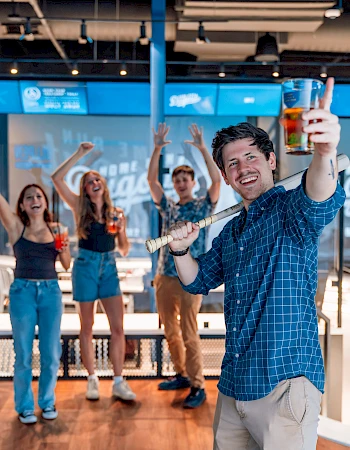 A group of people celebrating indoors. One person in front holds a drink and pool cue, while others cheer with raised glasses in the background.