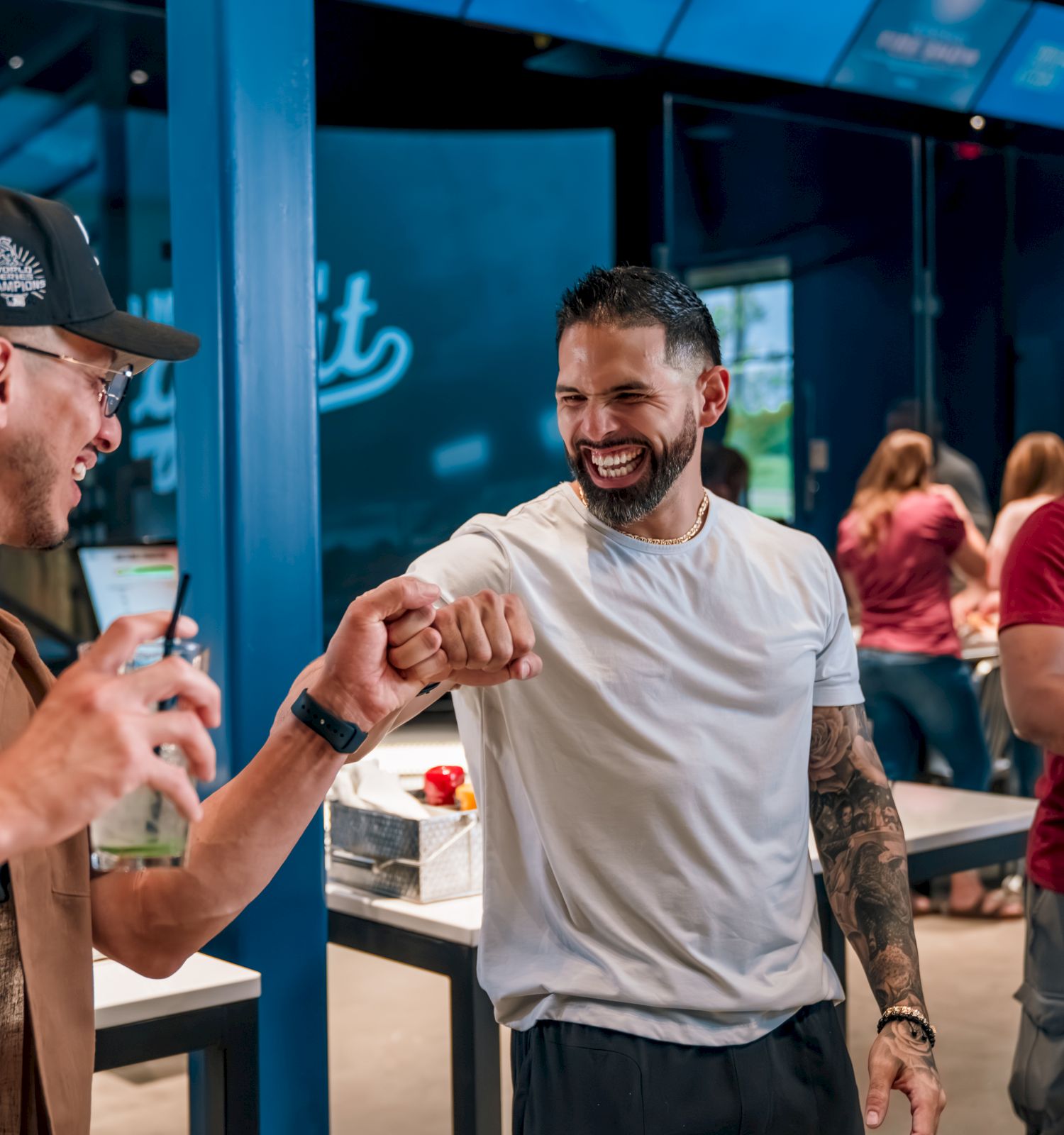 Three men in a modern cafe or lounge share a friendly fist bump, smiling and chatting as others mingle in the background.