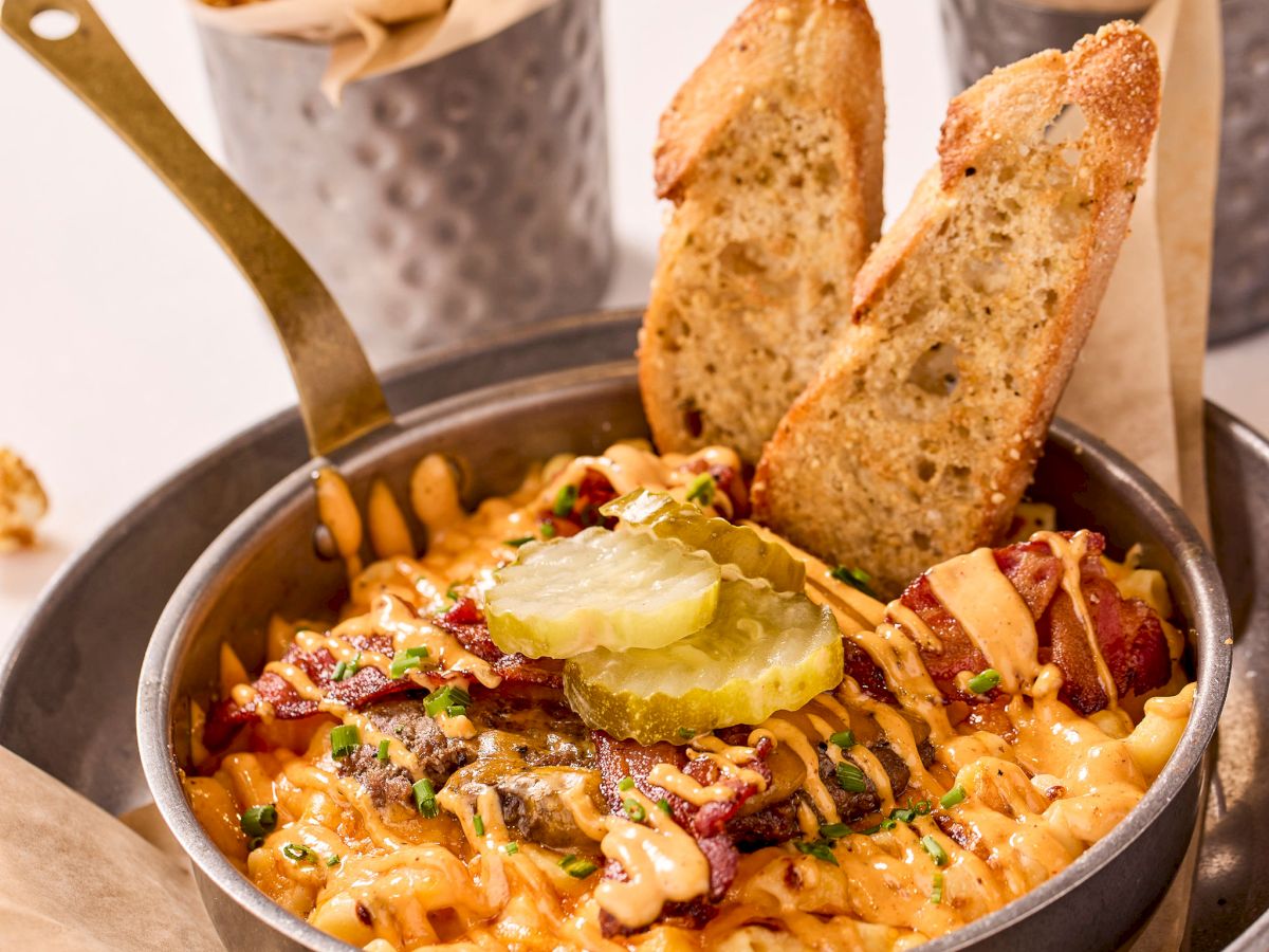 A skillet of loaded fries topped with pickles, sauce, and herbs, accompanied by toasted bread slices; popcorn and peanuts in the background.