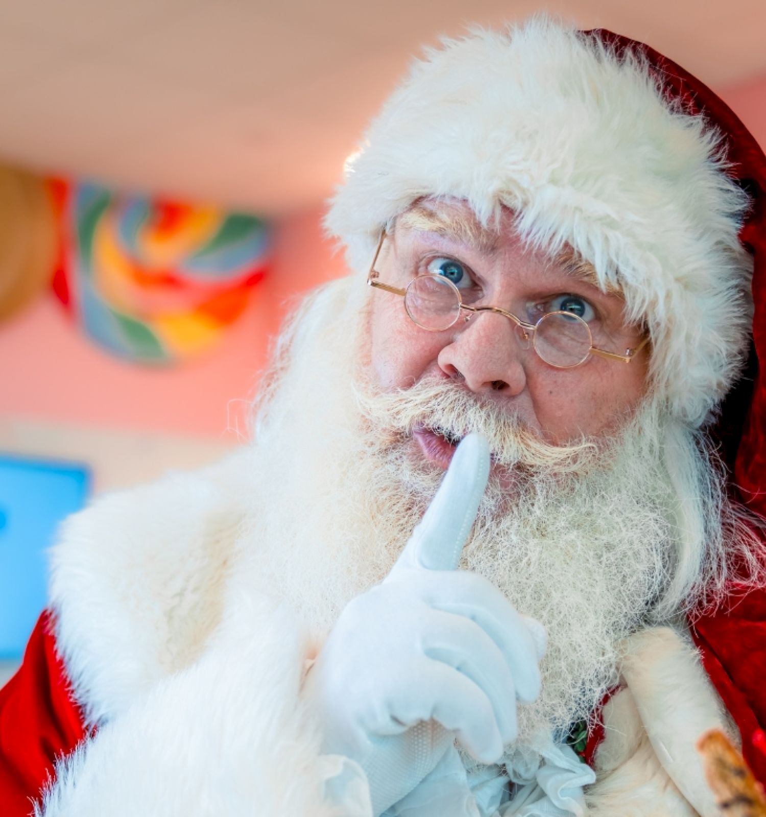 A person dressed as Santa Claus is holding a finger to their lips as a gesture to be quiet, in a festive setting with candy decor.
