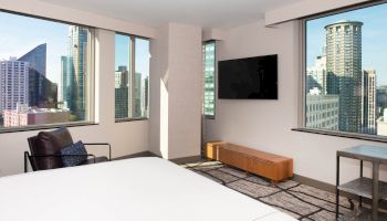 A modern hotel room features large windows, a bed, wall-mounted TV, chair, and cityscape views with tall buildings.