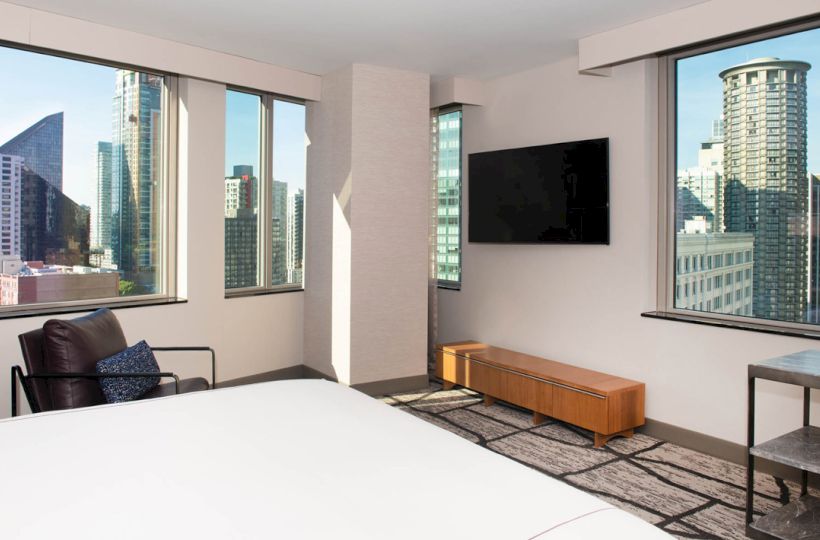A modern hotel room features large windows, a bed, wall-mounted TV, chair, and cityscape views with tall buildings.