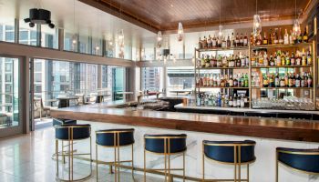 A modern bar with high stools, shelves stocked with bottles, hanging lights, and a view of city buildings through large windows.
