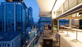 A modern city rooftop terrace with seating overlooks a cityscape of high-rise buildings, captured at dusk with illuminated interiors.
