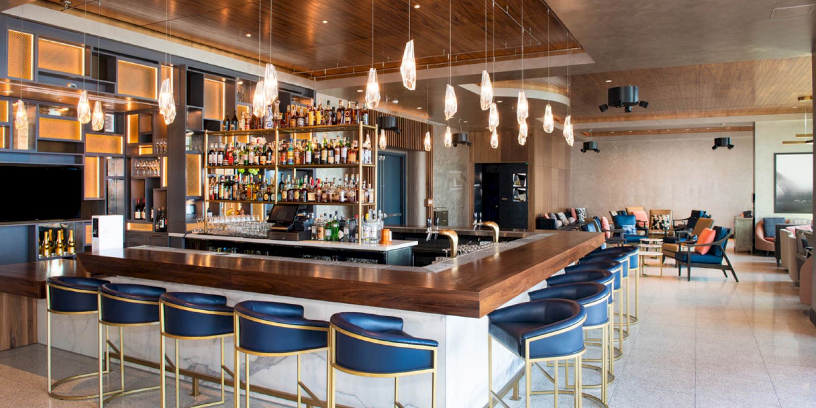 A modern bar with a wooden counter, blue barstools, and hanging pendant lights, featuring a well-stocked shelf of liquor bottles.
