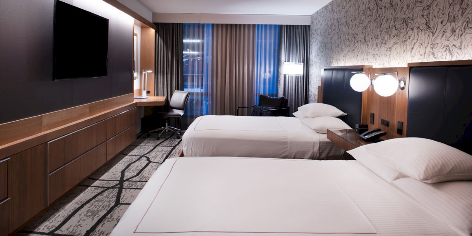 A hotel room with two beds, a TV, work desk, lamps, and modern decor.