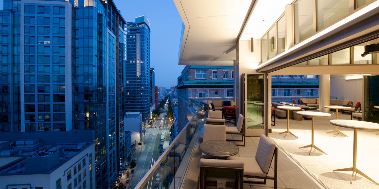 A modern cityscape at dusk with tall buildings, viewed from a stylish rooftop terrace with tables and chairs.