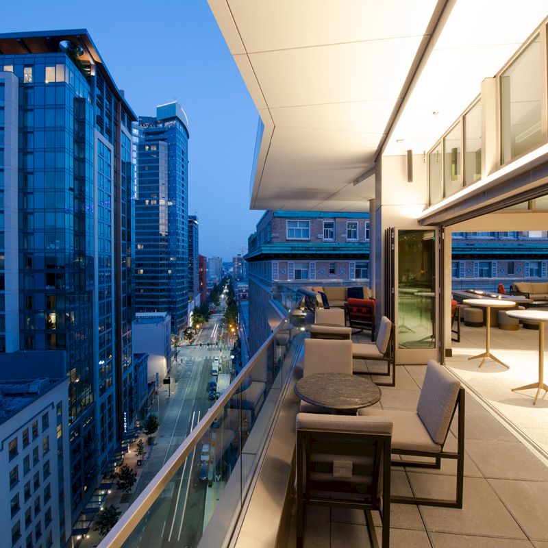 A modern cityscape at dusk with a view from a balcony featuring casual seating and illuminated buildings along a street.