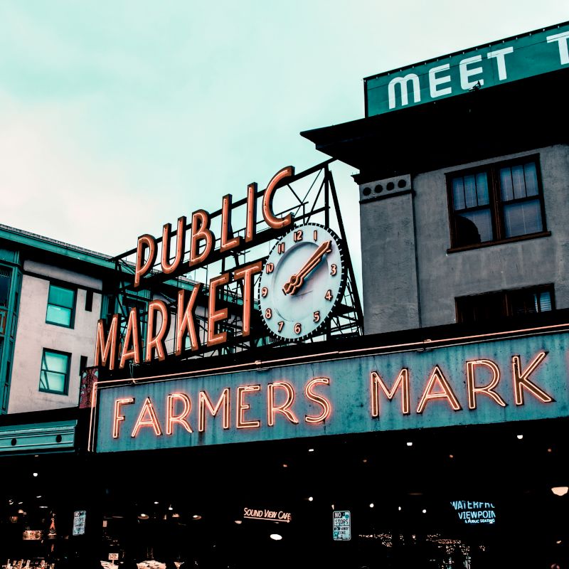 A historic public market sign with a clock and neon "Farmer's Market" text on a building's facade, suggesting a vibrant urban setting.
