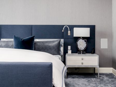 A modern bedroom with a navy blue headboard, white bedding, blue pillows, and a nightstand featuring a lamp and decorative items.