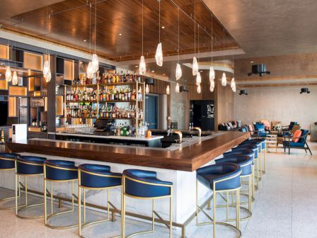 A modern bar with a wooden counter, blue stools, pendant lights, and a well-stocked liquor display, set in a spacious, stylish interior.