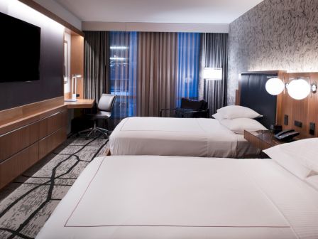 The image shows a modern hotel room with two beds, a TV, a desk with a chair, and stylish lighting and decor.