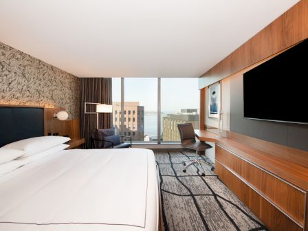 A modern hotel room features a large bed, a desk with a chair, a TV, and a window with a city view.