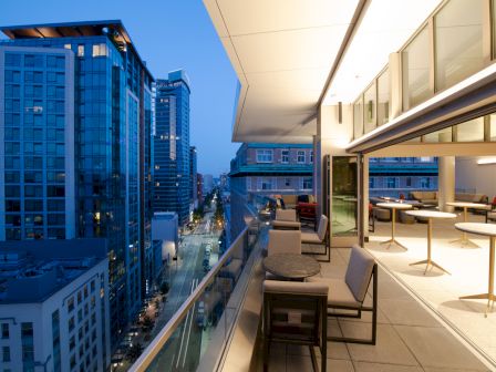 The image shows a modern urban balcony with outdoor seating, overlooking illuminated city skyscrapers at dusk.