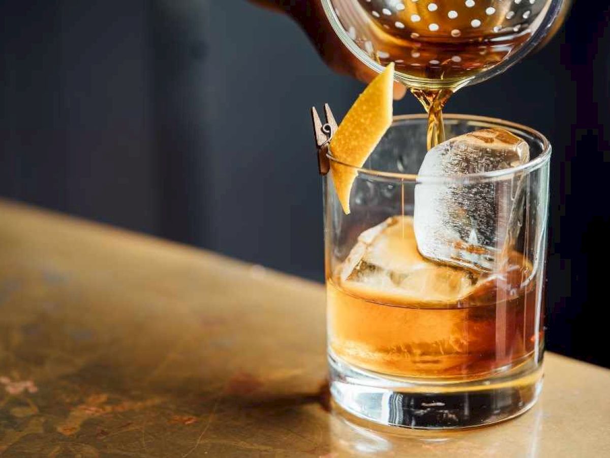 A cocktail is being poured into a glass with a large ice cube and a citrus peel garnish, set on a wooden surface.