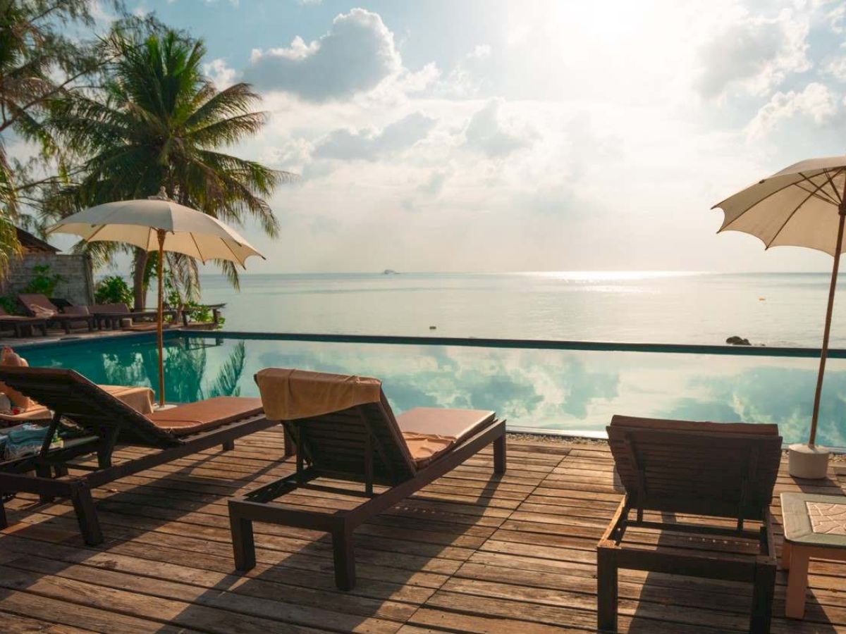 A serene beachfront with lounge chairs, umbrellas, and a view of the ocean. Palm trees and a sunny sky complete the tranquil scene.