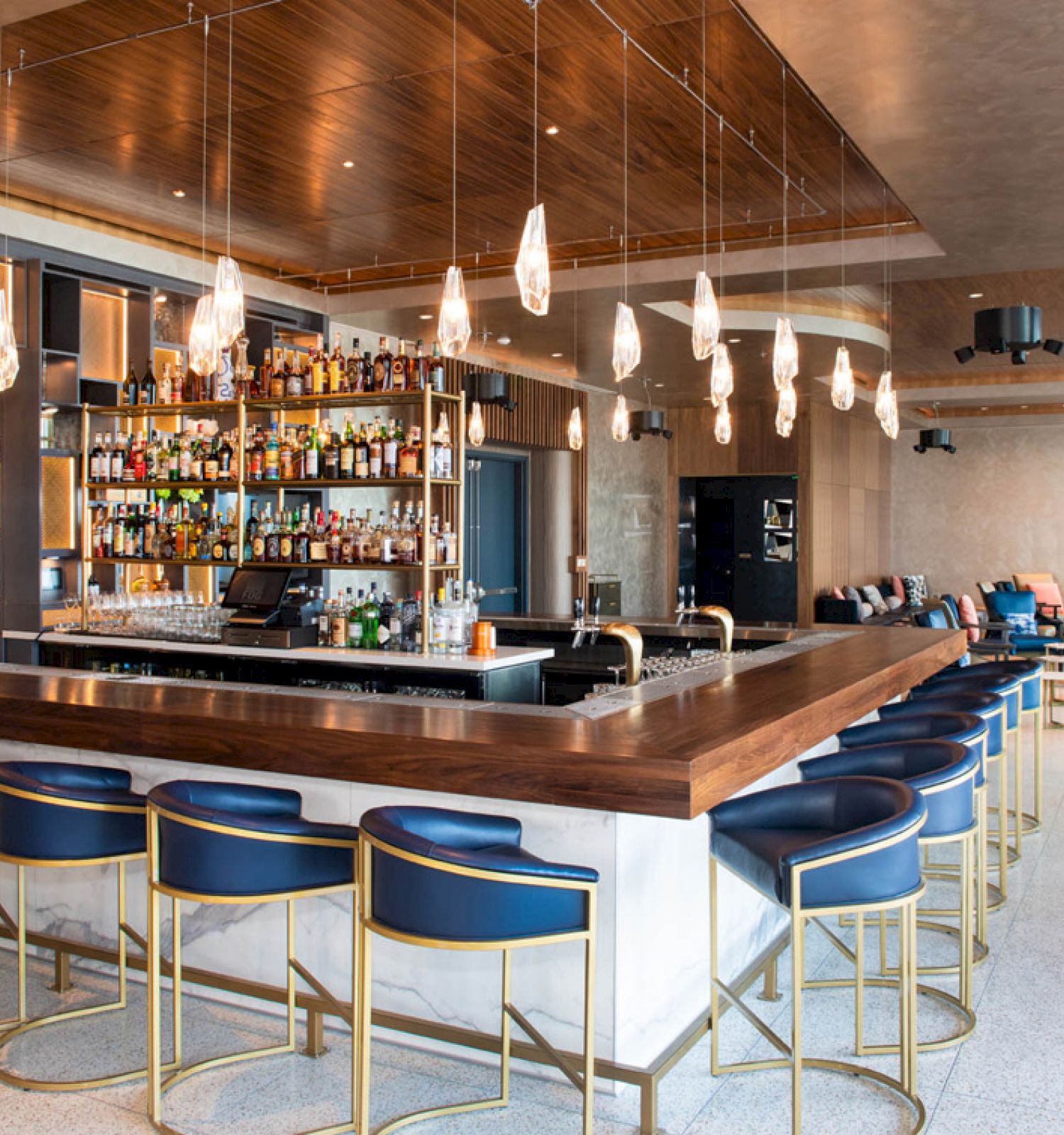 A modern bar with a stocked counter, blue stools, and hanging lights, located in a stylish lounge area with a mix of seating options.
