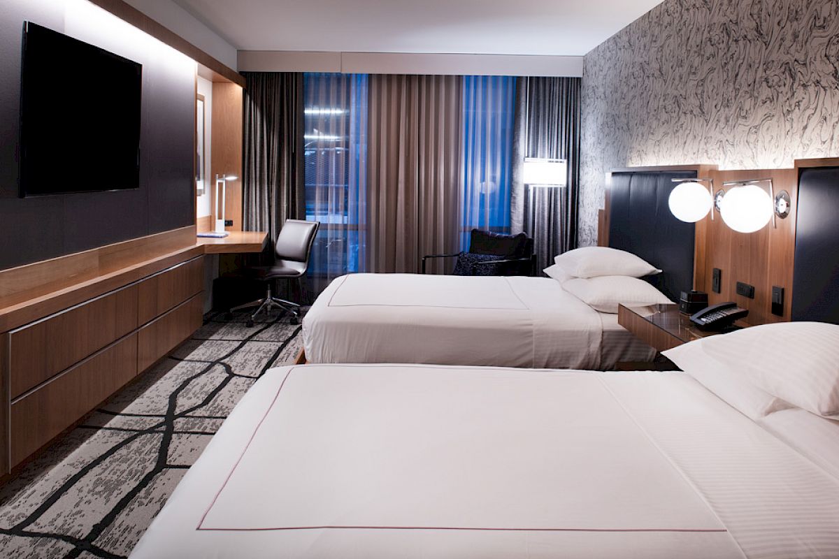 A modern hotel room with two beds, a flat-screen TV, a desk with a chair, and stylish lighting and decor, creating a cozy atmosphere.