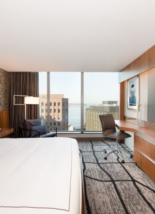 A modern hotel room with a large bed, wall-mounted TV, desk, chair, and window view of buildings and water.
