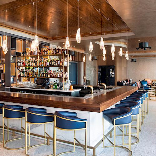 A modern bar with a central counter, blue upholstered stools, hanging lights, and shelves stocked with liquor bottles, in a stylish lounge.