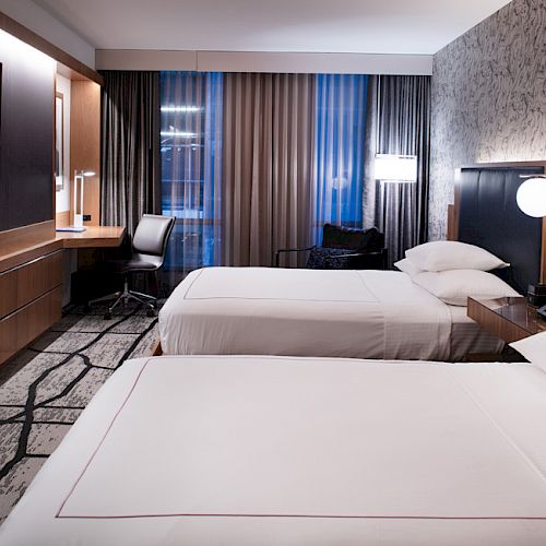 The image shows a modern hotel room with two beds, a large TV, a desk and chair, stylish lighting, and a patterned carpet and wall design.