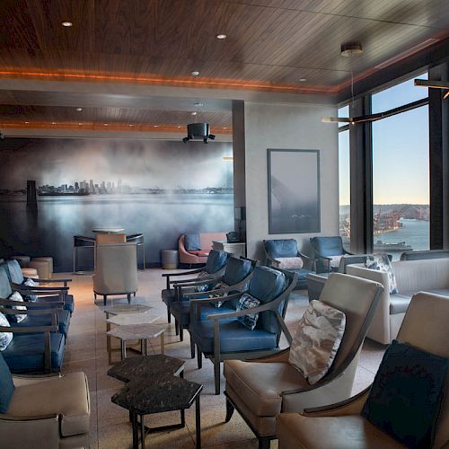 A modern lounge area with comfortable seating and large windows offering a scenic view of a waterfront and distant skyline.