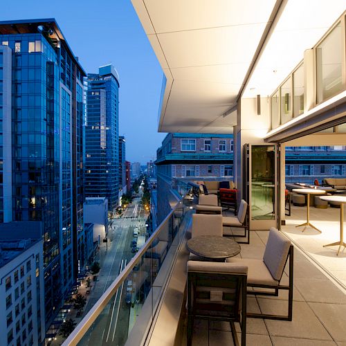 A modern cityscape from a high-rise balcony at dusk, with outdoor seating and nearby tall buildings lining a street below.