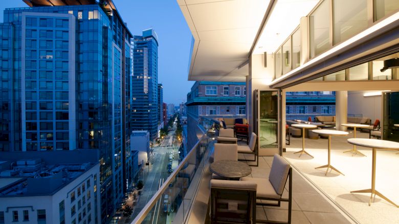 A modern cityscape view from a high balcony with outdoor seating and tables, overlooking illuminated skyscrapers at dusk.
