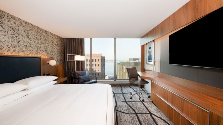 A modern hotel room with a large bed, desk, and TV, featuring a cityscape view through a window.