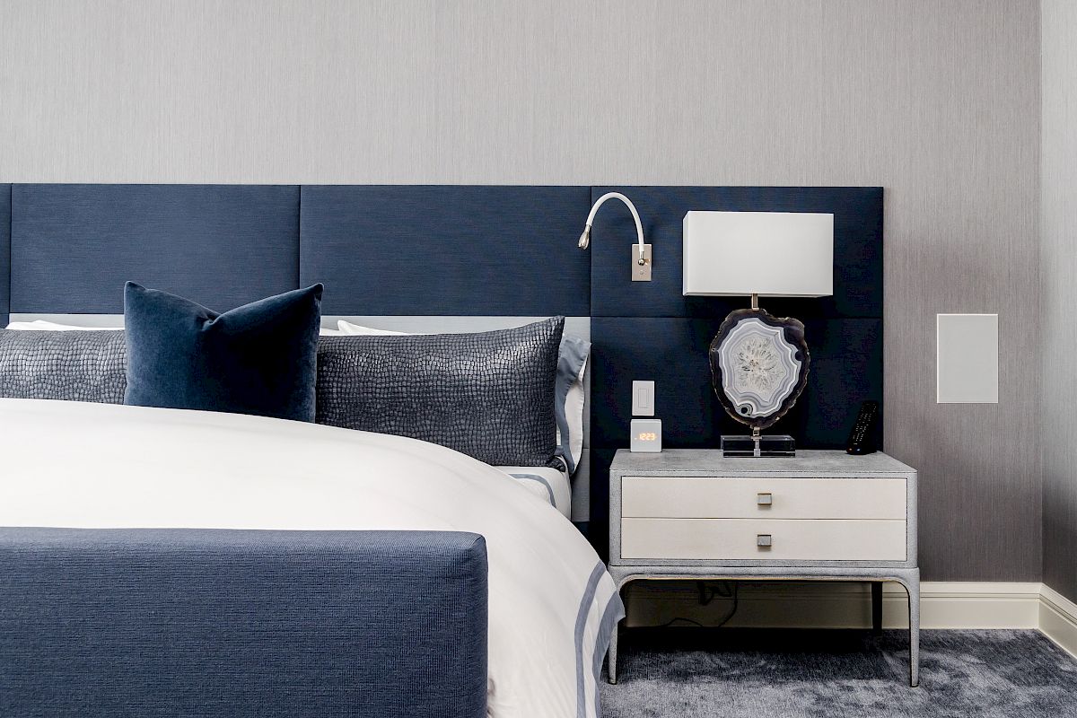 The image shows a modern bedroom with a blue and white color scheme, featuring a bed, pillows, nightstand, lamp, and stylish decor elements.