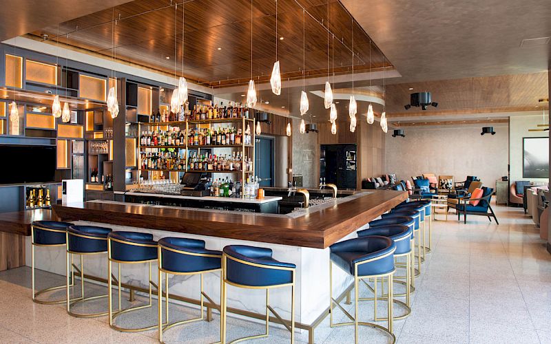 Chic bar with hanging lights, a variety of spirits on shelves, and blue cushioned bar stools creating a modern, inviting atmosphere.