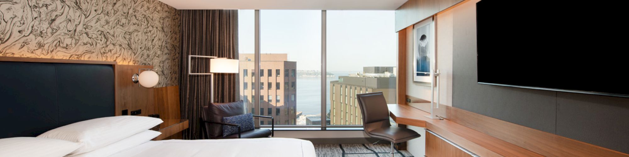 A modern hotel room with a bed, desk, chair, large TV, and window view of buildings and water in the background.